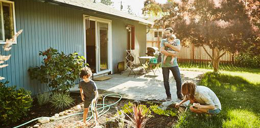 Little Kid watering plants with a house, while dad watches holding a baby and mom is weeding