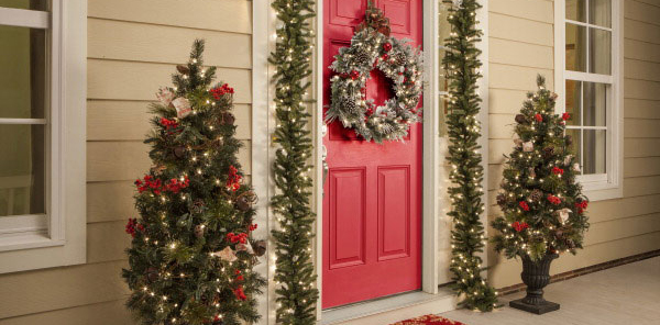 Red door decorated with wreaths and lights with two mini pine trees decorated next to the door