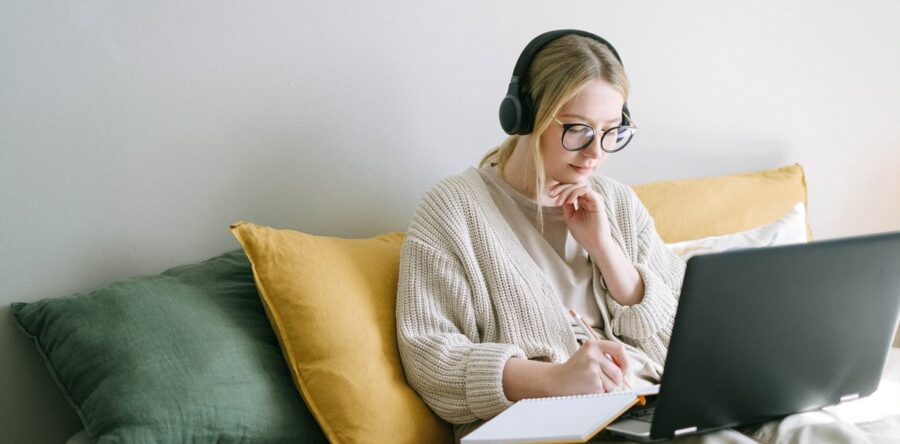 Woman writing on a notepad while looking at a laptop and wearing headphones