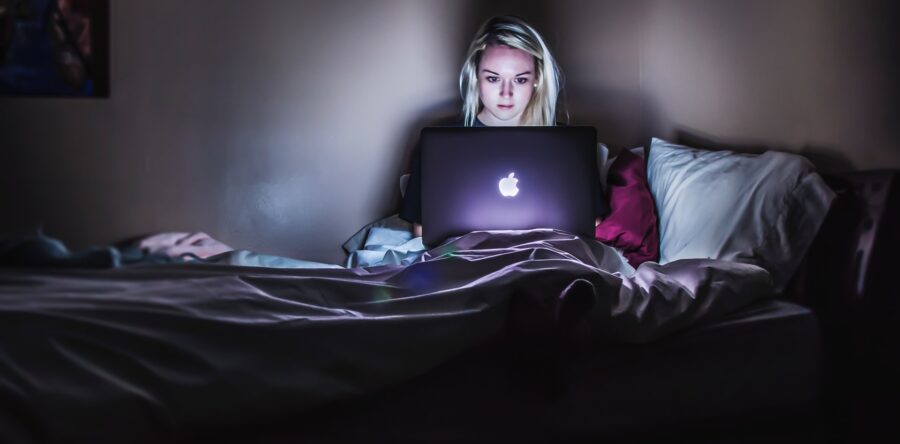Girl sitting on a bed in the dark with her laptop. The light from the laptop luminates her face