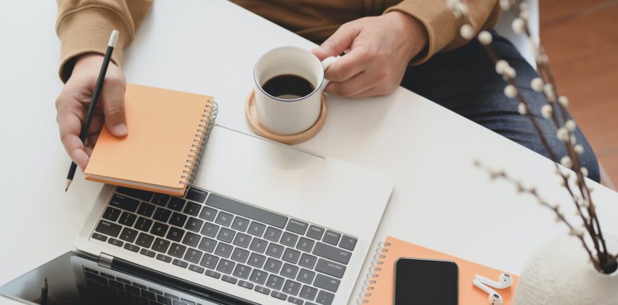 Person sitting at desk with notebooks, phone, mug of coffee, and laptop